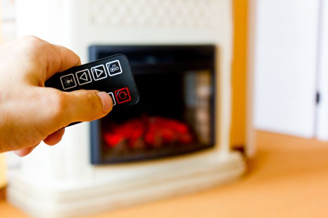 This is a stock photo of an electric fireplace with a hand holding a remote in the foreground.